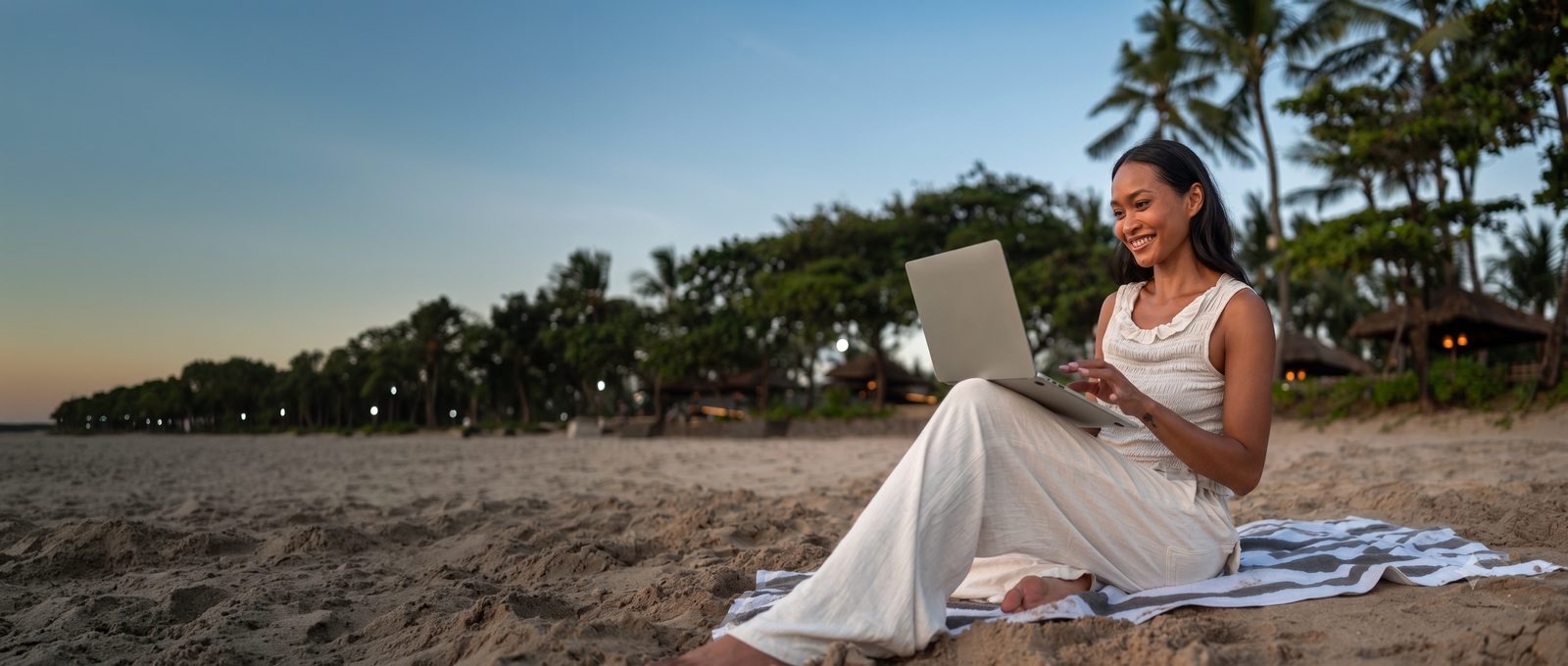 A woman sits on a beach towel at dusk with a laptop on her lap, smiling, palms and the shore behind her