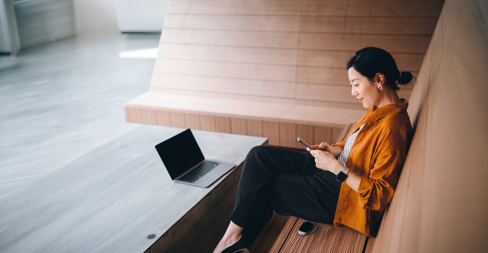 A woman in an orange shirt sits on wide wooden steps, checking her phone, with a laptop open on the step beside her